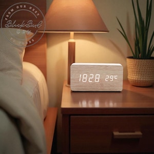 May include: A rectangular wooden digital clock displaying the time 18:28 and the temperature 29 degrees. The clock sits on a dark wooden nightstand next to a lamp and a potted plant.
