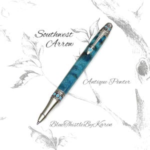 May include: A ballpoint pen with a shimmering teal blue barrel and antique pewter metal accents. The pen features a clip shaped like an arrow, inlaid with small turquoise-colored stones, and the text Southwest Arrow, Antique Pewter, and BlueThistleByKaren.
