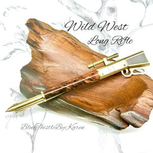 May include: A Wild West Long Rifle pen with a brown and gold finish. The pen has a wood-grain barrel and a gold-colored metal rifle design. The pen is displayed on a piece of wood.