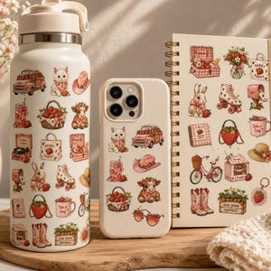 Pu&ograve; includere: Una borraccia, una custodia per telefono e un taccuino a spirale color crema decorati con adesivi a tema fragola. Gli adesivi presentano illustrazioni di fragole, animali e altri oggetti correlati. Gli articoli sono disposti su una superficie di legno.