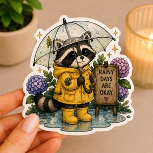 May include: A sticker featuring a cartoon raccoon wearing a yellow raincoat and boots, holding an umbrella. The raccoon stands in a puddle with a sign that reads "Rainy Days Are Okay." Purple flowers and stars surround the raccoon.
