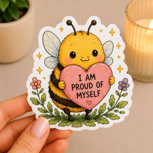 Può includere: Un adesivo con un'ape dei cartoni animati che tiene in mano un cuore rosa con la scritta "I AM PROUD OF MYSELF". L'ape è gialla e nera con le ali, circondata da stelle, fiori e fogliame verde. L'adesivo ha un bordo bianco.