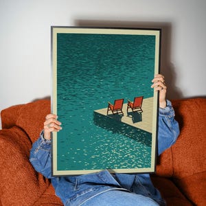 May include: A framed art print depicting a serene waterscape with two red lounge chairs on a dock. The water is a deep teal, and the sky is not visible. The print is held by a person wearing a denim jacket.