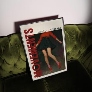 May include: A framed magazine cover titled "MOVEMENTS" with "ISSUE 01 - COURAGE" in red text. The cover art features a person in a red top and dark skirt. The magazine is resting on a green velvet sofa.