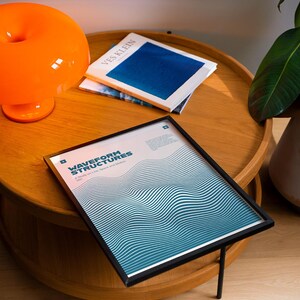 May include: A framed print with the text "WAVEFORM STRUCTURES" and a blue and white wave design, resting on a wooden table. An orange lamp and a book titled "VES KLEIN" are also on the table.