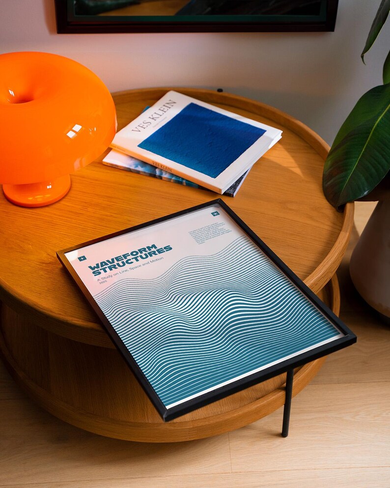 May include: A framed print with the text "WAVEFORM STRUCTURES" and a blue and white wave design, resting on a wooden table. An orange lamp and a book titled "VES KLEIN" are also on the table.