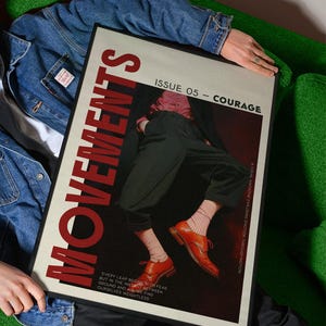 May include: A framed magazine cover with the title "MOVEMENTS" in large red letters. The cover features an image of a person wearing orange shoes and black pants. The text "ISSUE 05 - COURAGE" is also visible. The magazine is held by a person wearing a denim jacket.