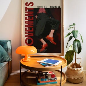 May include: A two-tiered wooden side table with black legs, topped with books and magazines. An orange mushroom-shaped lamp sits on the top tier. A framed poster with the word "MOVEMENTS" and the text "ISSUE 05 - COURAGE" hangs on the wall.