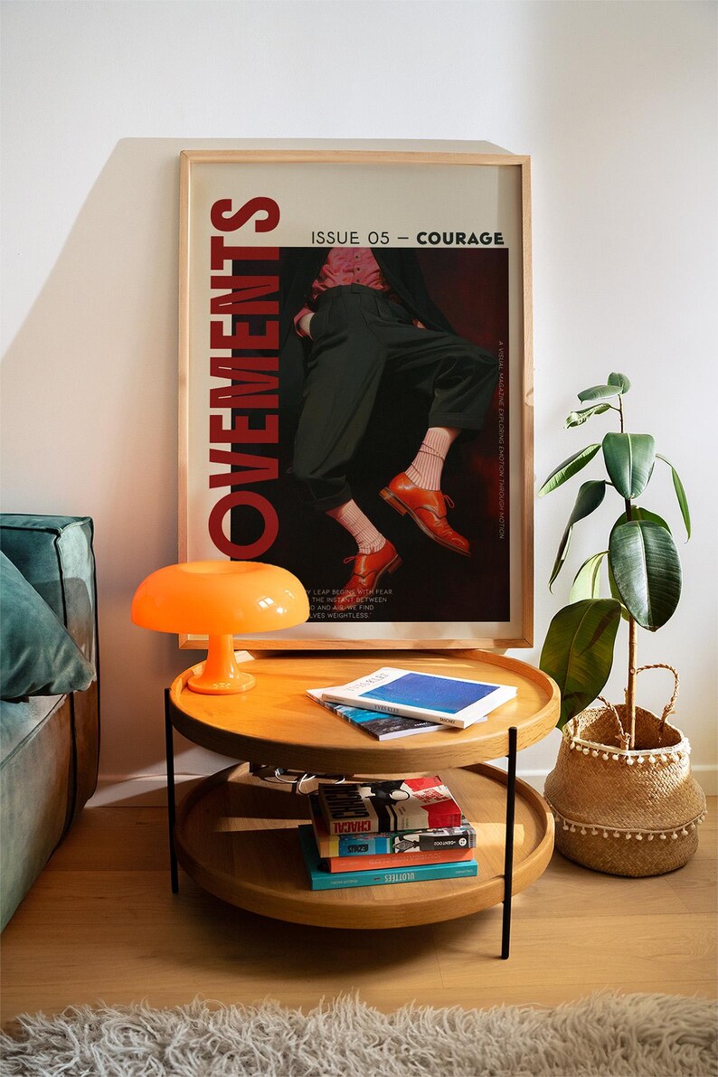 May include: A two-tiered wooden side table with black legs, topped with books and magazines. An orange mushroom-shaped lamp sits on the top tier. A framed poster with the word "MOVEMENTS" and the text "ISSUE 05 - COURAGE" hangs on the wall.