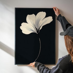 May include: A framed art print featuring a large, stylized cream-colored flower against a black background. The flower's petals are painted with broad strokes, and the stem is a thin, curved line. The artwork is held by a person.