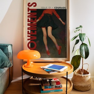 May include: A framed poster with the word "MOVEMENTS" in large red letters. The poster features a person in a red top and black skirt. A two-tiered wooden side table with books and an orange mushroom lamp sits in front of the poster.