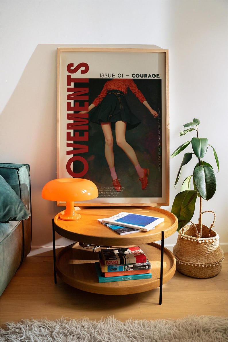 May include: A framed poster with the word "MOVEMENTS" in large red letters. The poster features a person in a red top and black skirt. A two-tiered wooden side table with books and an orange mushroom lamp sits in front of the poster.