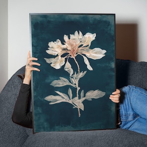 May include: A framed botanical print featuring a detailed illustration of a flower with large, delicate petals and leaves. The artwork is set against a dark teal background, creating a striking contrast. The print is encased in a black frame.