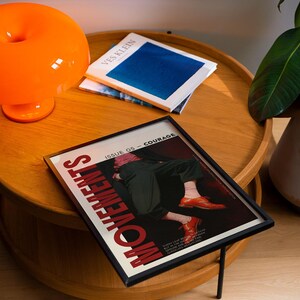 May include: A framed magazine cover with the word "MOVEMENTS" in red, featuring an illustration of a person in black pants and orange shoes. An orange lamp and two books are on a wooden table.