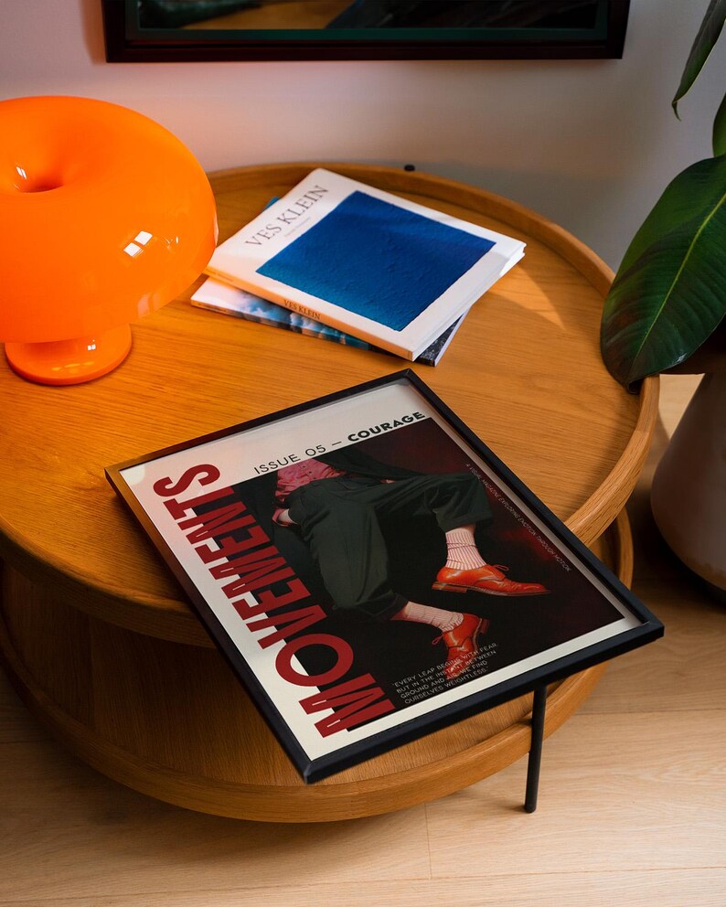 May include: A framed magazine cover with the word "MOVEMENTS" in red, featuring an illustration of a person in black pants and orange shoes. An orange lamp and two books are on a wooden table.