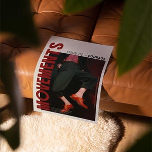 May include: A magazine titled "MOVEMENTS" with the text "ISSUE 05 - COURAGE" is displayed on a brown leather couch. The magazine cover features a person wearing dark green pants and orange shoes. A white fluffy rug is in the foreground.