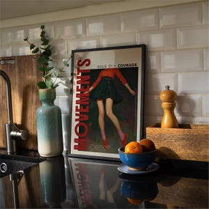 May include: A framed poster with the word "MOVEMENTS" in red, featuring a fashion illustration of a person in a red top and green skirt. The poster is on a black countertop with a blue bowl of oranges, a vase, and a wooden box.