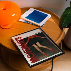 May include: A framed magazine cover with the word "MOVEMENTS" in red, featuring a person in a red top and black skirt. An orange lamp and books are on a wooden table. The magazine is titled "ISSUE 01 - COURAGE".