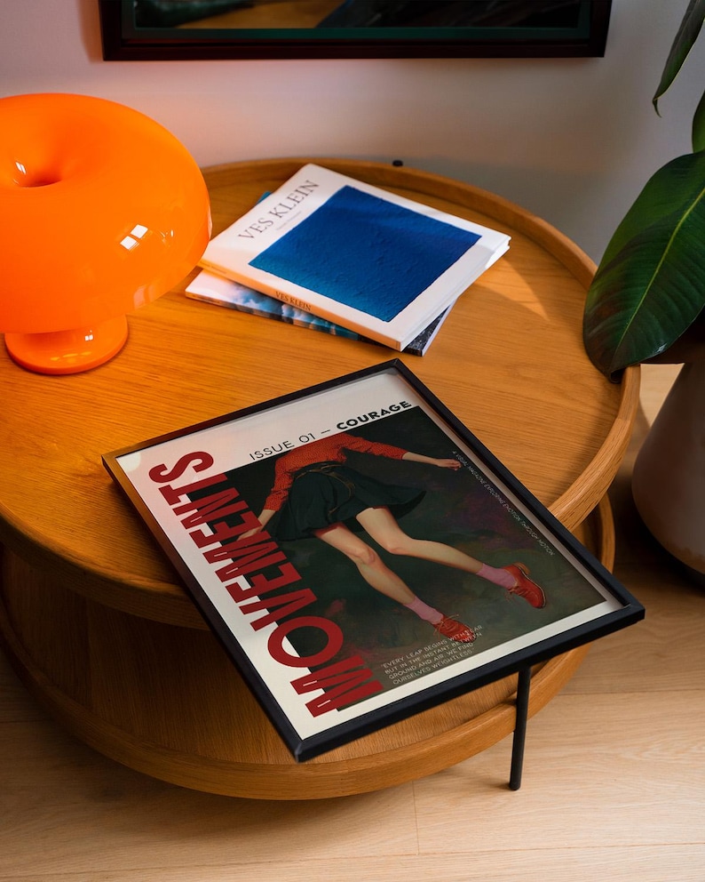 May include: A framed magazine cover with the word "MOVEMENTS" in red, featuring a person in a red top and black skirt. An orange lamp and books are on a wooden table. The magazine is titled "ISSUE 01 - COURAGE".