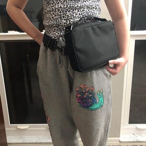 May include: A black lunch bag with a handle worn around the waist. The person is wearing a leopard print top and grey sweatpants. The sweatpants have a colourful snail graphic with mushrooms.