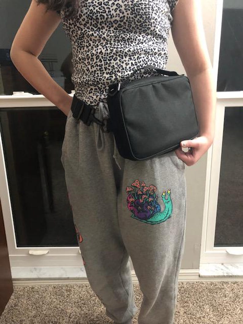 May include: A black lunch bag with a handle worn around the waist. The person is wearing a leopard print top and grey sweatpants. The sweatpants have a colourful snail graphic with mushrooms.