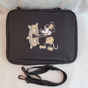 May include: Black rectangular travel bag featuring an embroidered image of Mickey Mouse holding a ship's wheel. The bag has a zipper closure and a detachable black shoulder strap. The Mickey Mouse image is in shades of black, yellow, and white.