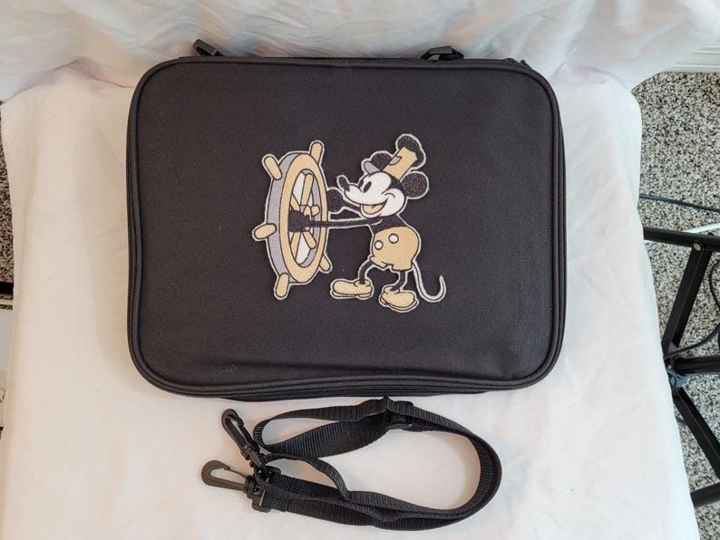May include: Black rectangular travel bag featuring an embroidered image of Mickey Mouse holding a ship's wheel. The bag has a zipper closure and a detachable black shoulder strap. The Mickey Mouse image is in shades of black, yellow, and white.