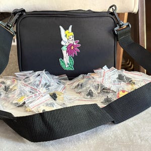 May include: Black crossbody bag with a Tinkerbell graphic and a collection of pins in small plastic bags. The bag has a black adjustable strap. The pins are of various colors and designs.