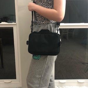 May include: A black rectangular crossbody bag with a shoulder strap. The bag is worn across the body. The bag has a handle on top. The person is wearing a leopard print top and grey sweatpants.