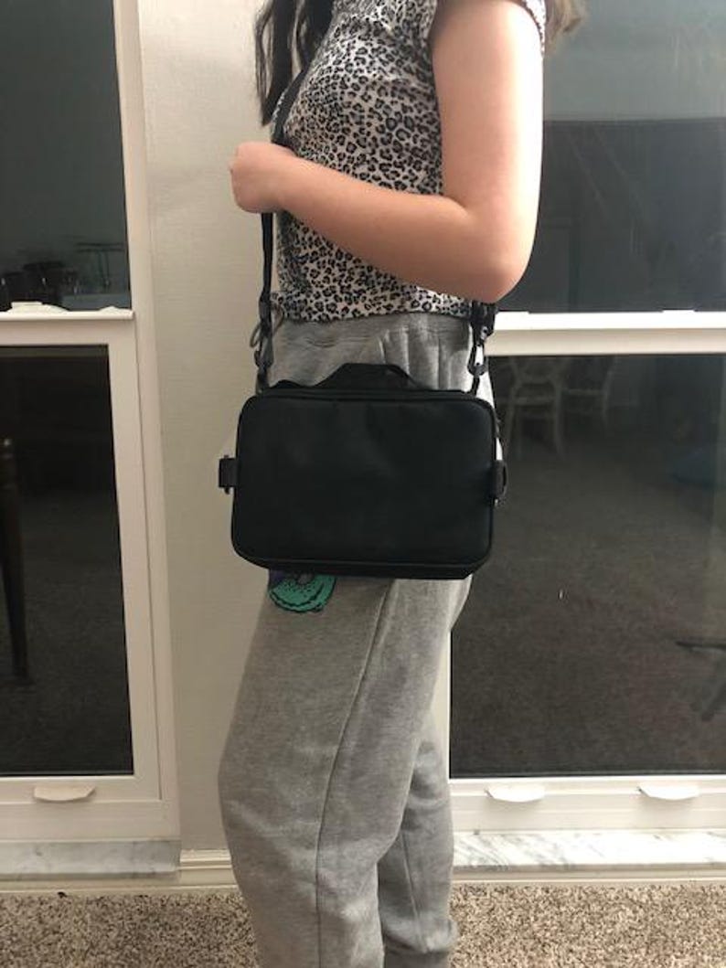 May include: A black rectangular crossbody bag with a shoulder strap. The bag is worn across the body. The bag has a handle on top. The person is wearing a leopard print top and grey sweatpants.