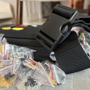 May include: Black fanny pack with a black buckle and strap. The fanny pack is filled with small, individually packaged pins. The pins are various colours and designs, including gold and silver.
