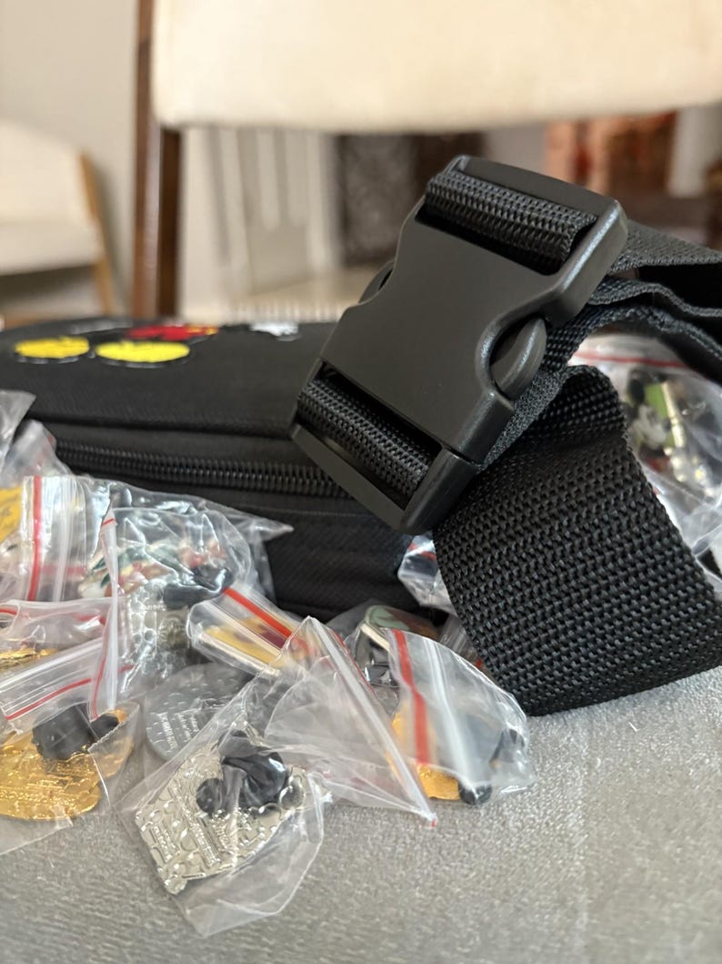 May include: Black fanny pack with a black buckle and strap. The fanny pack is filled with small, individually packaged pins. The pins are various colours and designs, including gold and silver.
