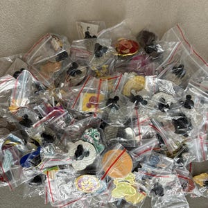 May include: A selection of enamel pins, each in a small, clear plastic bag with a red zip. The pins display diverse designs, including cartoon characters and abstract shapes. The pins are spread across a neutral surface. The pins are in individual bags.