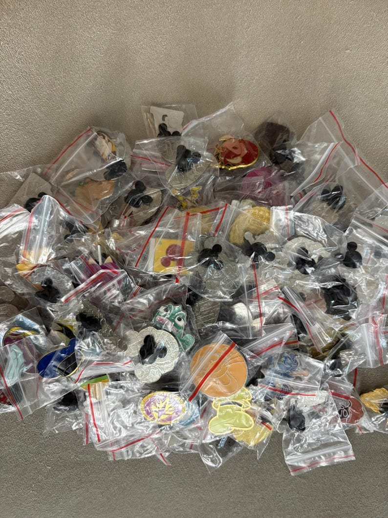 May include: A selection of enamel pins, each in a small, clear plastic bag with a red zip. The pins display diverse designs, including cartoon characters and abstract shapes. The pins are spread across a neutral surface. The pins are in individual bags.