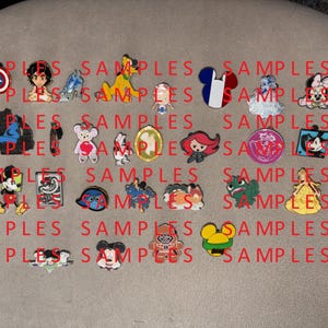May include: A selection of colourful enamel pins showcasing cartoon characters and symbols. Designs include a shield, a character with a blue outfit, a yellow dog, a French flag, a cloud, a bear with a heart, a bat, Mickey Mouse, and more. The word "SAMPLES" is printed in red.