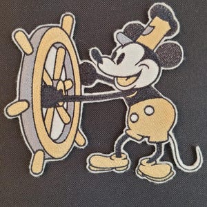 May include: Embroidered patch featuring Mickey Mouse in a captain's outfit, steering a ship's wheel. The design uses black, yellow, gray, and white thread. The patch has a white border and is likely for clothing or accessories.