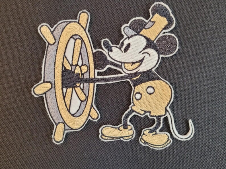 May include: Embroidered patch featuring Mickey Mouse in a captain's outfit, steering a ship's wheel. The design uses black, yellow, gray, and white thread. The patch has a white border and is likely for clothing or accessories.