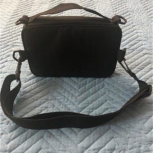 May include: A black camera bag with a shoulder strap and a top handle. The bag is rectangular with a zippered closure and metal clips for the strap. The bag is on a light blue quilted surface.