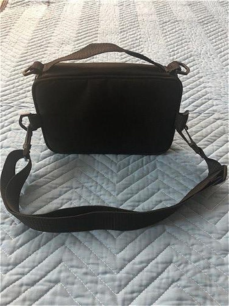 May include: A black camera bag with a shoulder strap and a top handle. The bag is rectangular with a zippered closure and metal clips for the strap. The bag is on a light blue quilted surface.