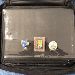 May include: Black zippered carrying case with clear plastic sleeves, holding three Disney pins. The pins feature cartoon characters and scenes, including a Sorcerer Mickey Mouse, a framed "Silly Symphonies" pin, and a castle design.