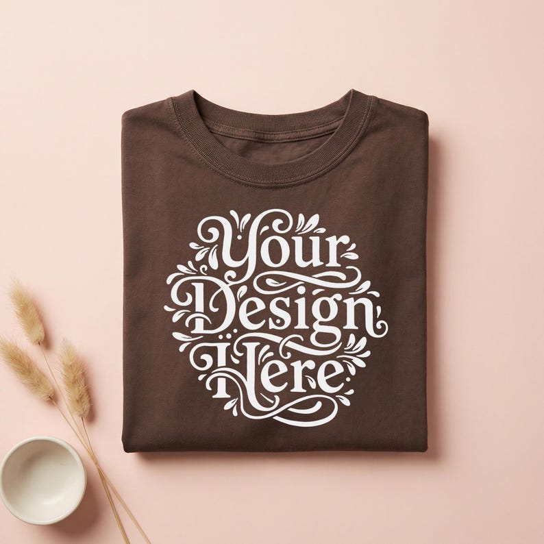 May include: A folded brown t-shirt with a white decorative design that reads "Your Design Here". The shirt is made of a soft material and is displayed on a light pink surface. A small white bowl and dried plants are in the lower left corner.