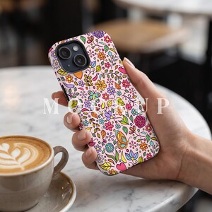 以下が含まれることがあります： 花、葉、蝶など、カラフルな花柄のピンク色の携帯電話ケース。 ケースは手に持たれており、手前にはコーヒーカップがあります。