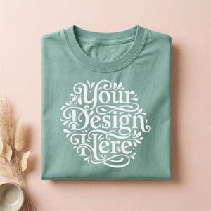 May include: A folded, seafoam green t-shirt with the text "Your Design Here" in a white, ornate script. The shirt is on a light pink surface, with dried plants and a small bowl in the lower left corner.