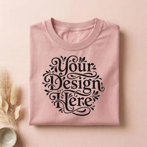 May include: A folded, light pink t-shirt with a crew neck. The shirt features a black, ornate design that reads "Your Design Here" in a circular arrangement. The shirt is made of a soft material.