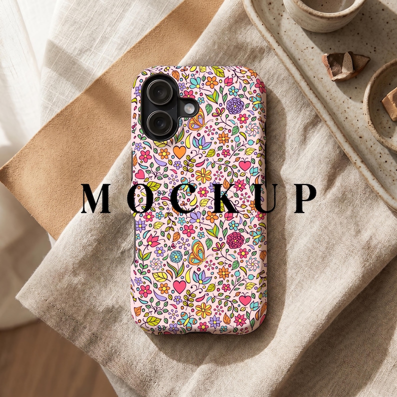 Peut inclure: Une coque de t&eacute;l&eacute;phone avec un motif floral color&eacute;, comprenant des fleurs, des c&oelig;urs et des papillons. Le fond de la coque est rose et le mot "MOCKUP" est imprim&eacute; en noir. La coque est pos&eacute;e sur une surface de couleur neutre.