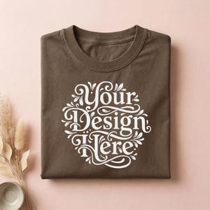 May include: A folded, brown t-shirt with a white, ornate design that reads "Your Design Here." The shirt has a crew neck and is made of a soft material. The design is centered on the front of the shirt.