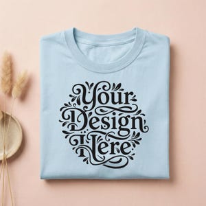 May include: A light blue t-shirt with a crew neck, folded neatly. The shirt features a black, ornate design in the center with the words "Your Design Here" in a decorative font. The shirt is on a light pink surface.