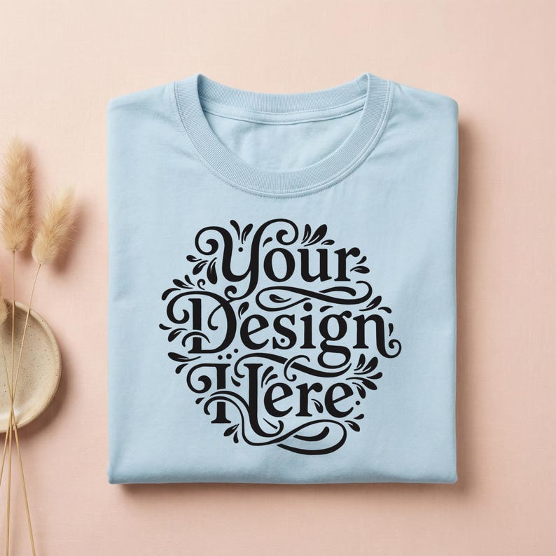 May include: A light blue t-shirt with a crew neck, folded neatly. The shirt features a black, ornate design in the center with the words "Your Design Here" in a decorative font. The shirt is on a light pink surface.