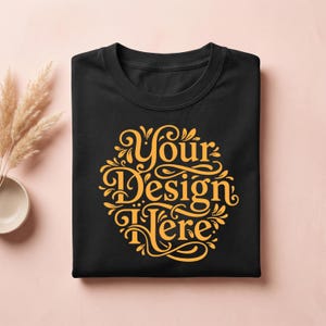 May include: Black crew neck t-shirt with a gold design that reads "Your Design Here" in an ornate, circular font. The shirt is neatly folded and presented on a light pink surface. Decorative elements are visible in the background.