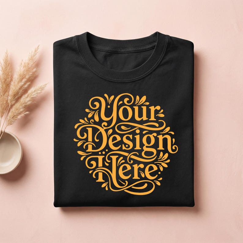 May include: Black crew neck t-shirt with a gold design that reads "Your Design Here" in an ornate, circular font. The shirt is neatly folded and presented on a light pink surface. Decorative elements are visible in the background.